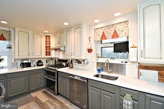 kitchen featuring a sink, light wood-type flooring, gray cabinets, under cabinet range hood, and appliances with stainless steel finishes