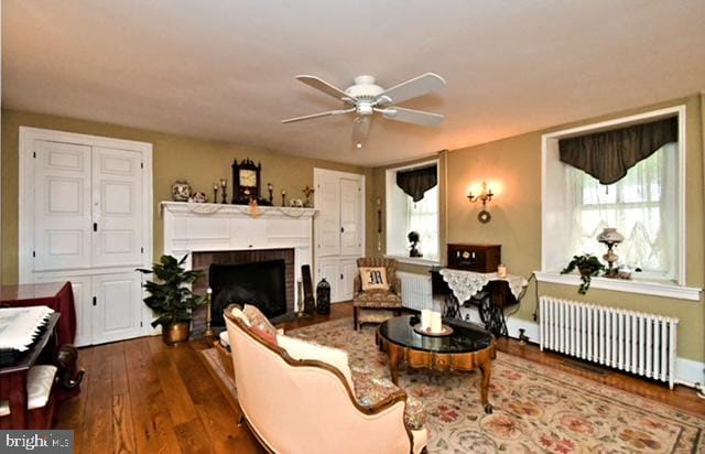 living room featuring radiator heating unit, a brick fireplace, ceiling fan, and dark wood-style flooring