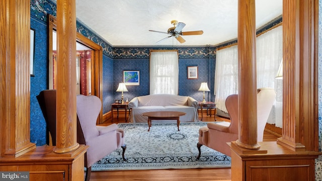 sitting room with a ceiling fan, wood finished floors, wallpapered walls, and decorative columns