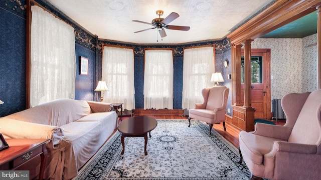 living area featuring decorative columns, wallpapered walls, wood finished floors, ceiling fan, and crown molding