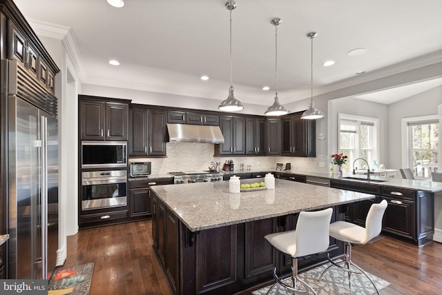 kitchen with built in appliances, a sink, dark wood finished floors, under cabinet range hood, and a kitchen island