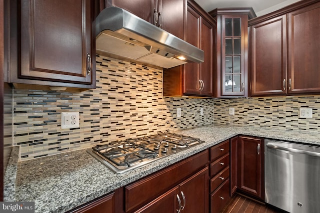 kitchen featuring stainless steel appliances, under cabinet range hood, backsplash, light stone countertops, and glass insert cabinets