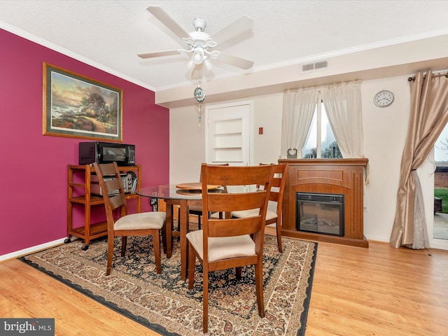 dining space with a ceiling fan, a glass covered fireplace, ornamental molding, visible vents, and wood finished floors