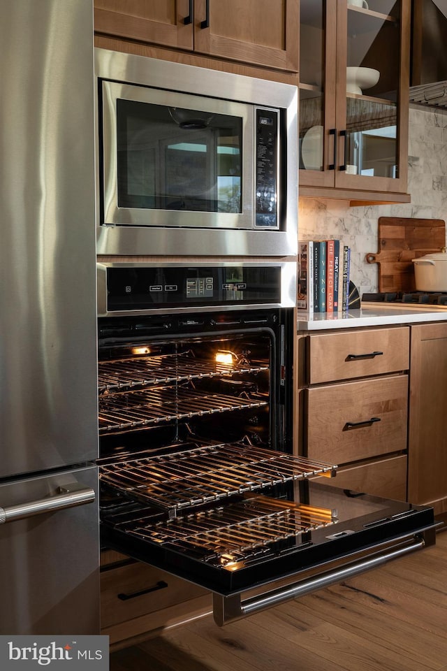 interior space with glass insert cabinets, light countertops, appliances with stainless steel finishes, wood finished floors, and backsplash