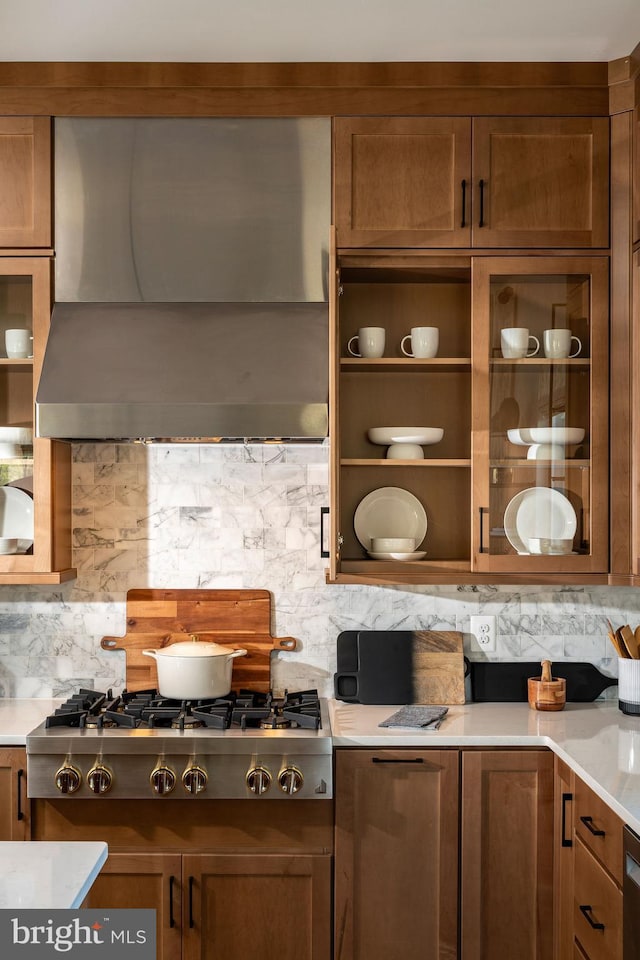 interior space featuring wall chimney exhaust hood, glass insert cabinets, stainless steel gas cooktop, light countertops, and backsplash