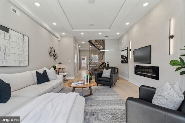 living room with a tray ceiling, light wood-type flooring, visible vents, recessed lighting, and stairs