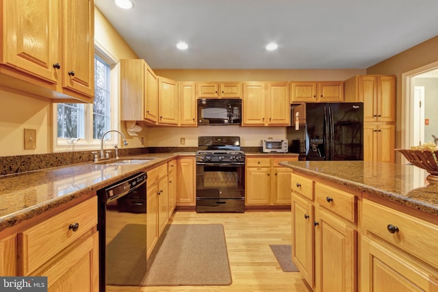 kitchen with light brown cabinets, dark stone countertops, black appliances, light wood finished floors, and a sink