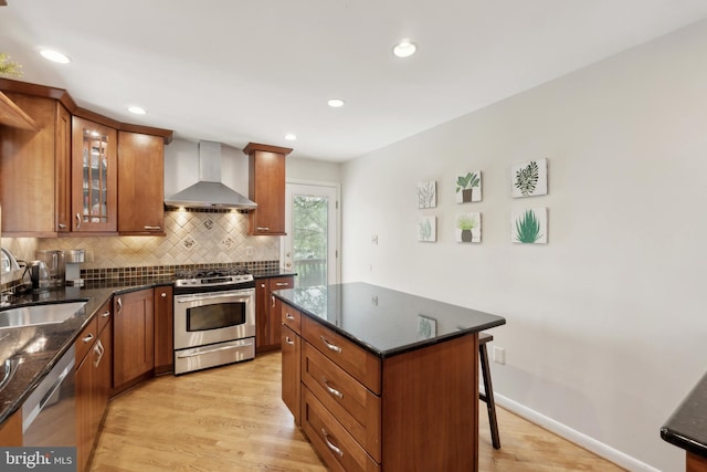 kitchen featuring wall chimney range hood, brown cabinets, a sink, stainless steel appliances, and light wood finished floors