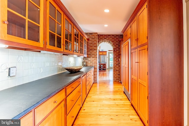 kitchen with light wood finished floors, decorative backsplash, brick wall, glass insert cabinets, and dark countertops
