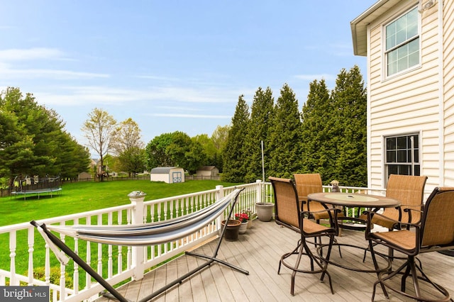 deck with outdoor dining area, a trampoline, an outdoor structure, a yard, and a storage unit