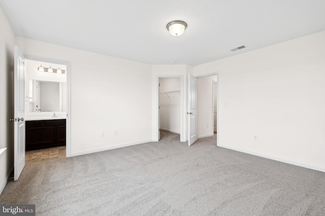 unfurnished bedroom featuring ensuite bathroom, visible vents, light carpet, a walk in closet, and baseboards