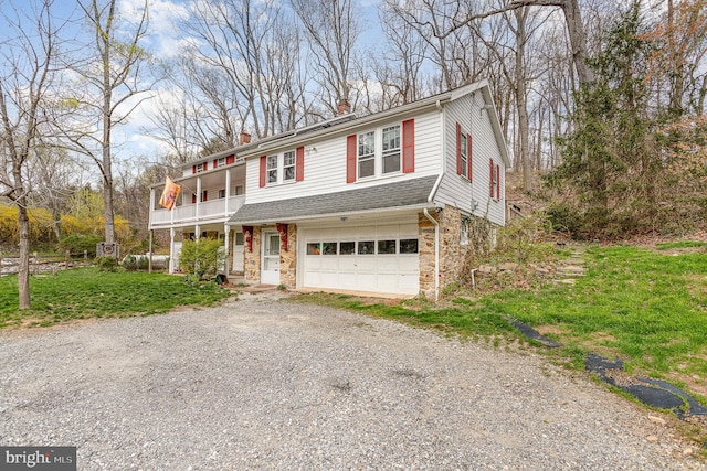 traditional-style house with a balcony, a chimney, stone siding, an attached garage, and gravel driveway