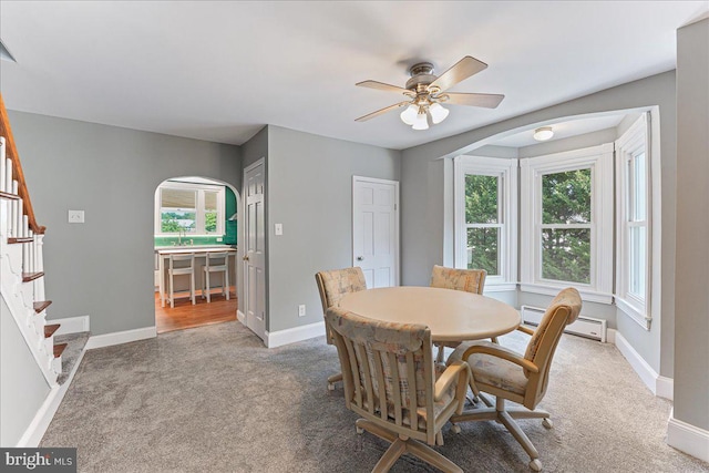 carpeted dining space featuring baseboards, arched walkways, a baseboard radiator, a ceiling fan, and stairs