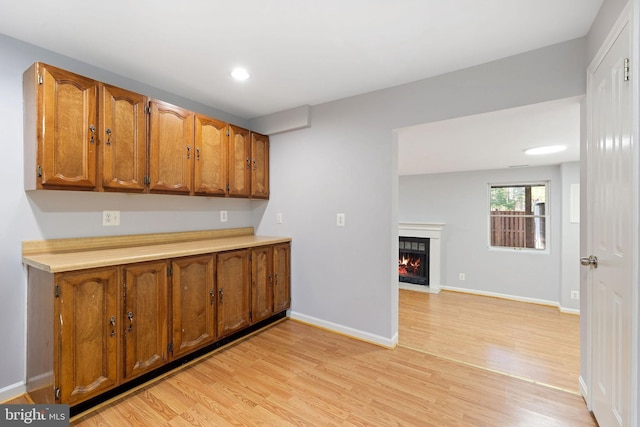 kitchen featuring light wood-style floors, baseboards, brown cabinetry, and a warm lit fireplace