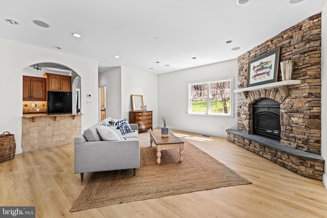 living area with baseboards, arched walkways, a stone fireplace, recessed lighting, and light wood-style flooring