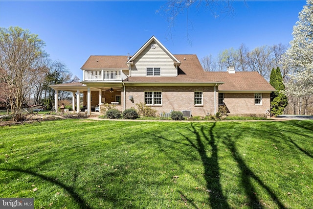 back of house featuring a balcony, a chimney, a ceiling fan, brick siding, and a lawn
