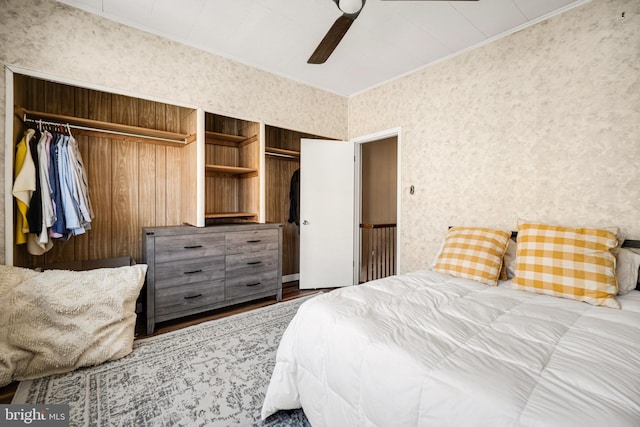 bedroom with crown molding, two closets, ceiling fan, wood finished floors, and wallpapered walls