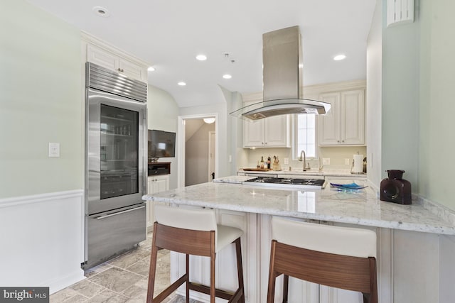 kitchen with light stone counters, island range hood, a peninsula, white gas stovetop, and stainless steel built in refrigerator