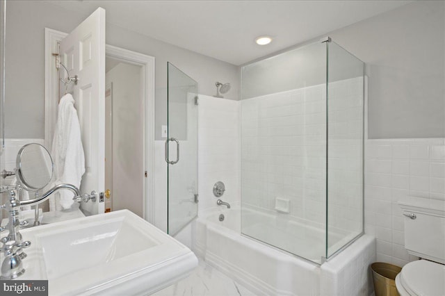 bathroom featuring toilet, marble finish floor, a sink, combined bath / shower with glass door, and tile walls