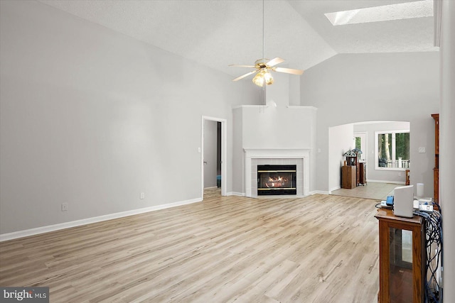 unfurnished living room featuring ceiling fan, a skylight, light wood-style floors, a tile fireplace, and high vaulted ceiling
