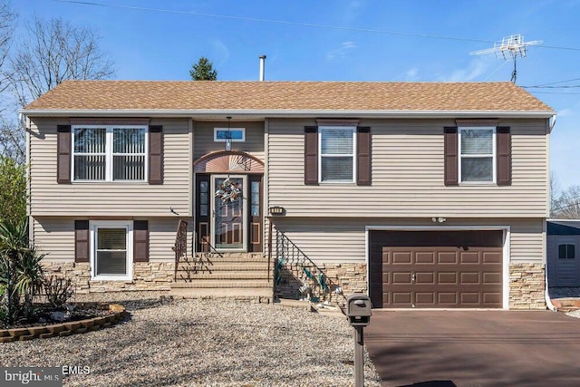 bi-level home featuring an attached garage, a shingled roof, driveway, and stone siding