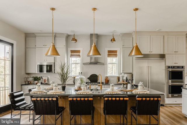 kitchen with wall chimney exhaust hood, light wood-type flooring, appliances with stainless steel finishes, a kitchen breakfast bar, and an island with sink