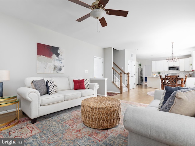 living room featuring light wood-type flooring, ceiling fan with notable chandelier, baseboards, and stairs
