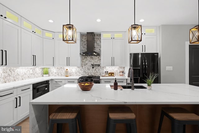 kitchen featuring wall chimney exhaust hood, black appliances, a sink, decorative backsplash, and an island with sink