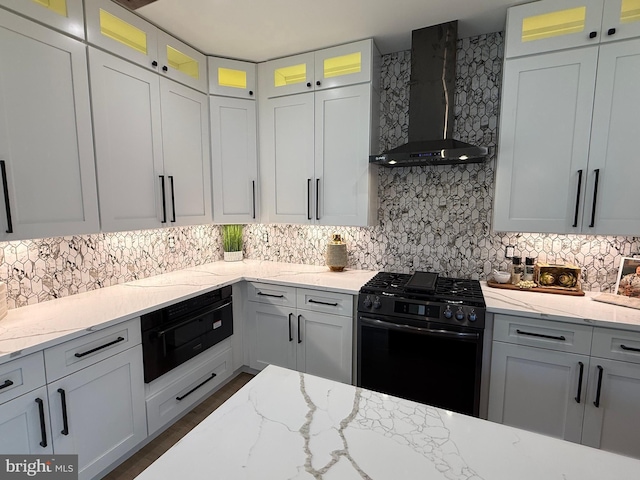 kitchen featuring black range with gas cooktop, wall chimney range hood, backsplash, light stone countertops, and glass insert cabinets