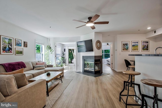 living room with ceiling fan, a multi sided fireplace, light wood-style flooring, baseboards, and bar