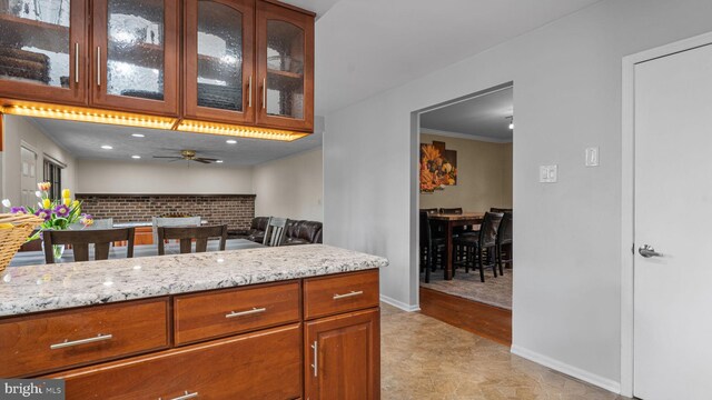 kitchen featuring brown cabinetry, light stone counters, a ceiling fan, and glass insert cabinets