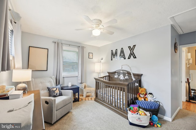 carpeted bedroom featuring a crib, baseboards, attic access, and ceiling fan