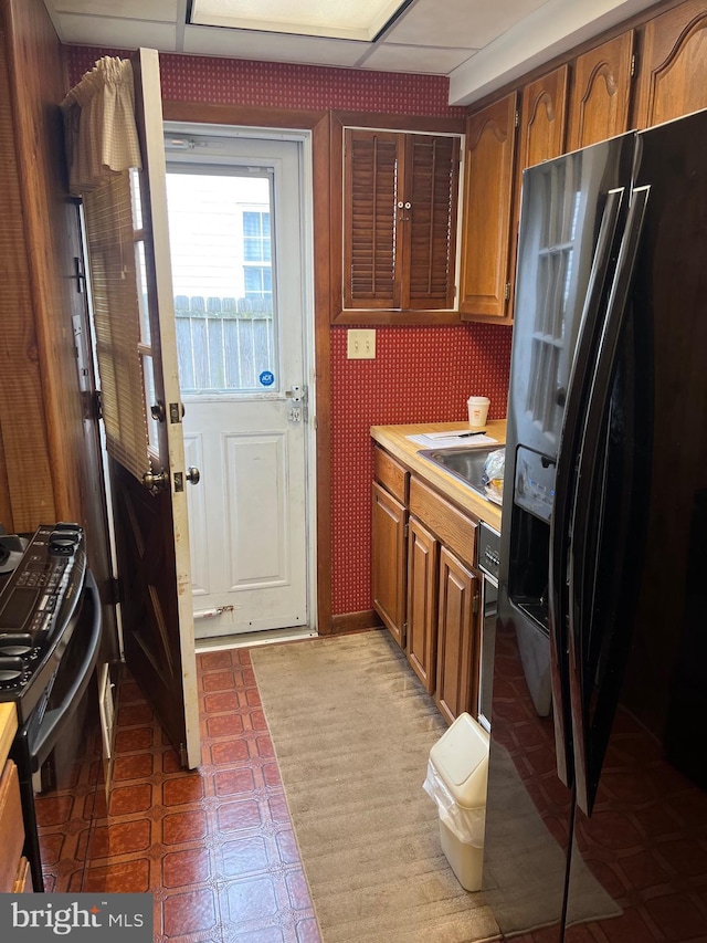 kitchen featuring brown cabinetry, black appliances, a sink, light countertops, and wallpapered walls