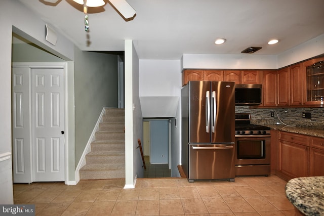 kitchen featuring glass insert cabinets, ceiling fan, dark stone counters, appliances with stainless steel finishes, and light tile patterned floors