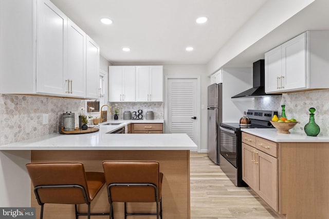 kitchen featuring wall chimney exhaust hood, a peninsula, light wood finished floors, appliances with stainless steel finishes, and a breakfast bar area