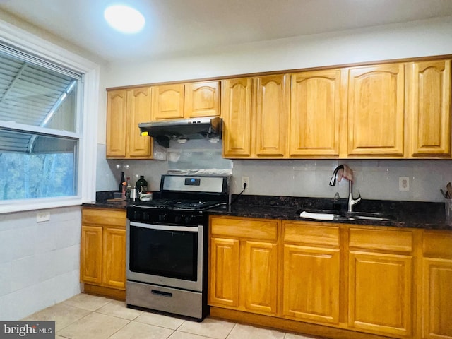 kitchen featuring dark stone countertops, light tile patterned floors, stainless steel gas range oven, under cabinet range hood, and a sink