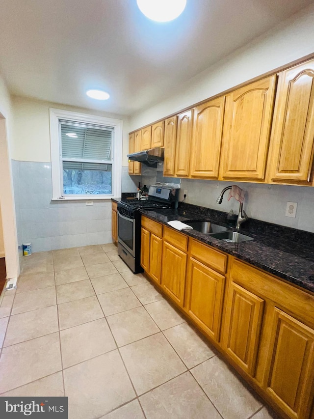 kitchen with dark stone countertops, light tile patterned floors, a sink, under cabinet range hood, and gas stove