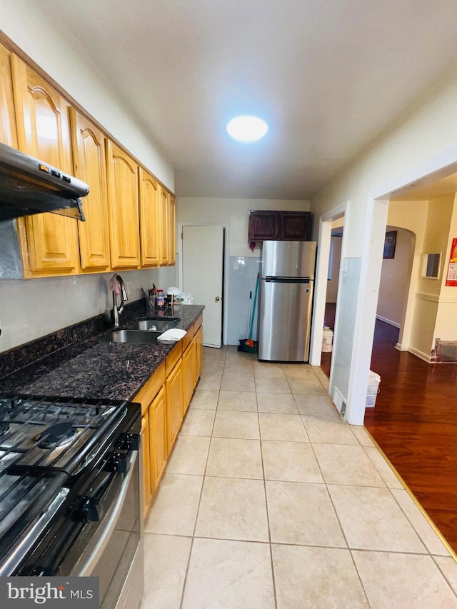 kitchen featuring light tile patterned floors, dark stone countertops, appliances with stainless steel finishes, under cabinet range hood, and a sink