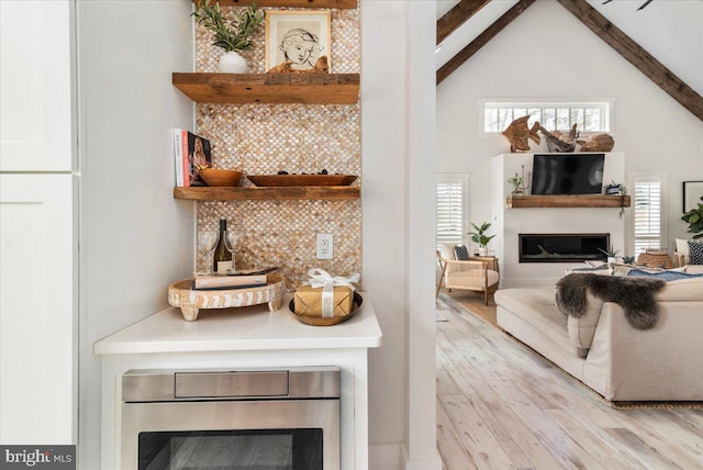 interior space featuring a glass covered fireplace, beam ceiling, decorative backsplash, high vaulted ceiling, and light wood finished floors