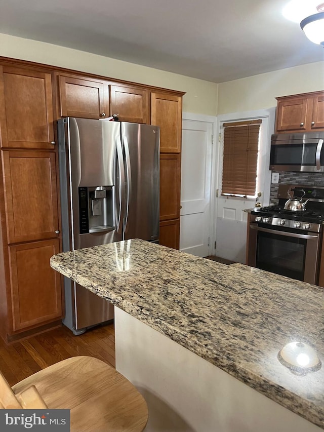 kitchen with light stone countertops, stainless steel appliances, brown cabinetry, and dark wood-type flooring