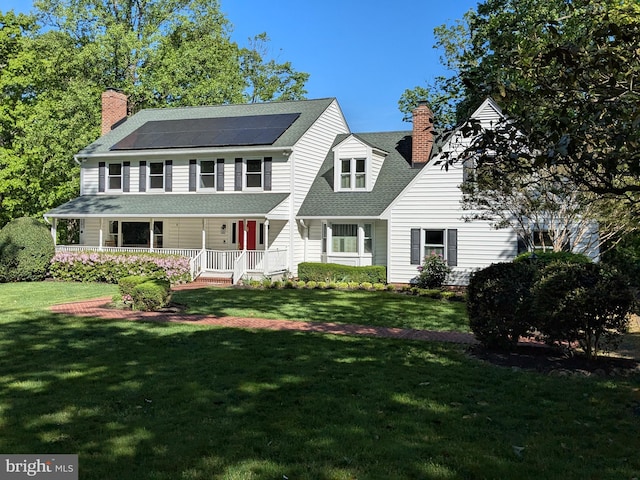 view of front of property with a chimney, a porch, roof mounted solar panels, a shingled roof, and a front yard