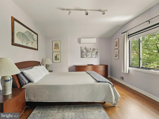 bedroom featuring visible vents, a wall mounted AC, rail lighting, wood finished floors, and baseboards
