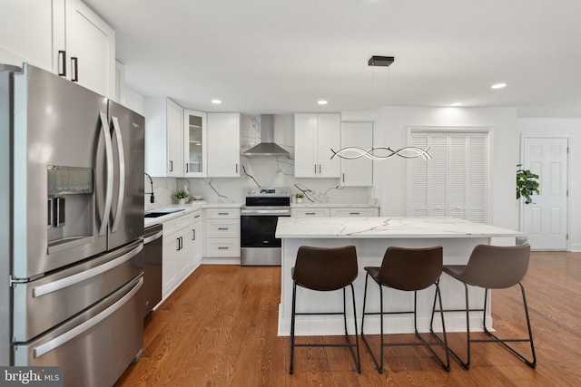 kitchen with wall chimney range hood, a sink, glass insert cabinets, appliances with stainless steel finishes, and a breakfast bar area