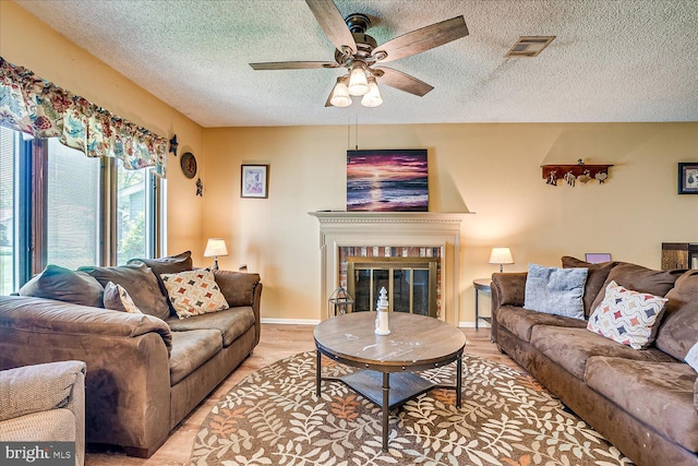 living room featuring visible vents, a glass covered fireplace, light wood-style floors, ceiling fan, and a textured ceiling