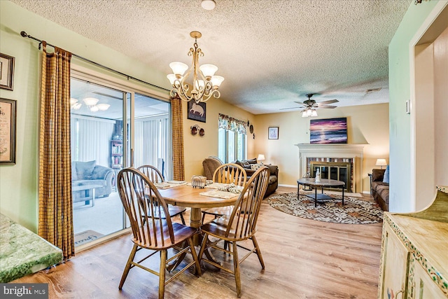 dining area with a textured ceiling, light wood finished floors, ceiling fan with notable chandelier, and a glass covered fireplace