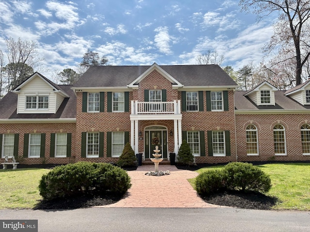 georgian-style home featuring roof with shingles, a balcony, a front lawn, and brick siding