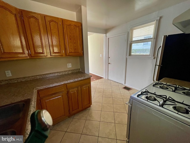 5714 63rd Ave, Riverdale, MD 20737 - kitchen with white gas range, freestanding refrigerator, brown cabinetry, light tile patterned flooring, and wall chimney exhaust hood