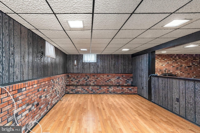 finished basement featuring wood walls, light wood finished floors, brick wall, and a paneled ceiling