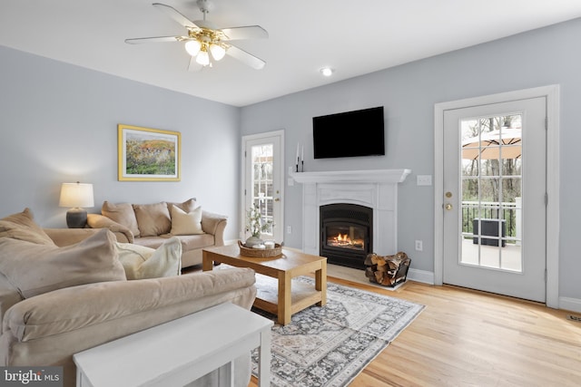 living area with a glass covered fireplace, a ceiling fan, light wood-style flooring, and baseboards