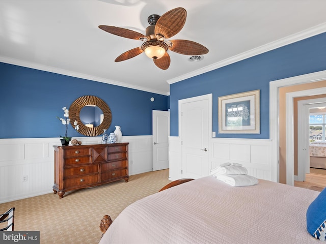 carpeted bedroom featuring crown molding, a wainscoted wall, visible vents, ceiling fan, and a decorative wall